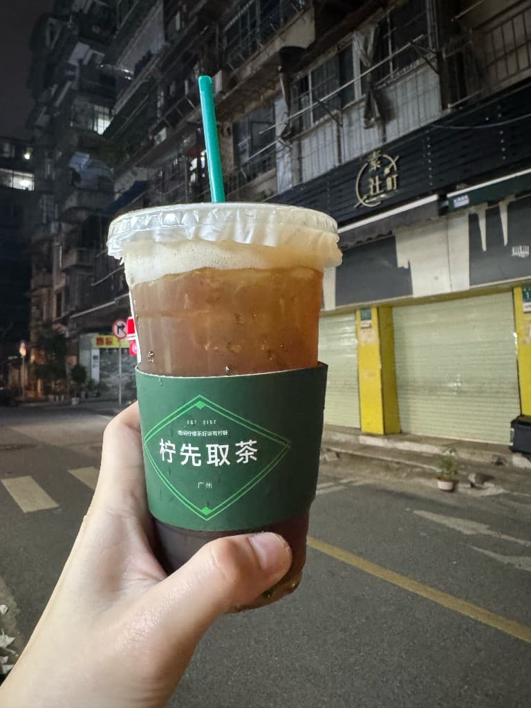 2026 年夜晚街头，手持柠先取茶，杯套印着「广州」二字，背景是熟悉的街景与斑马线